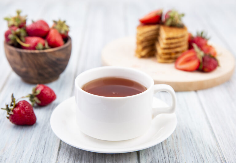 side view of cup of tea on saucer and waffle biscuits with strawberries in plate and bowl on wooden background